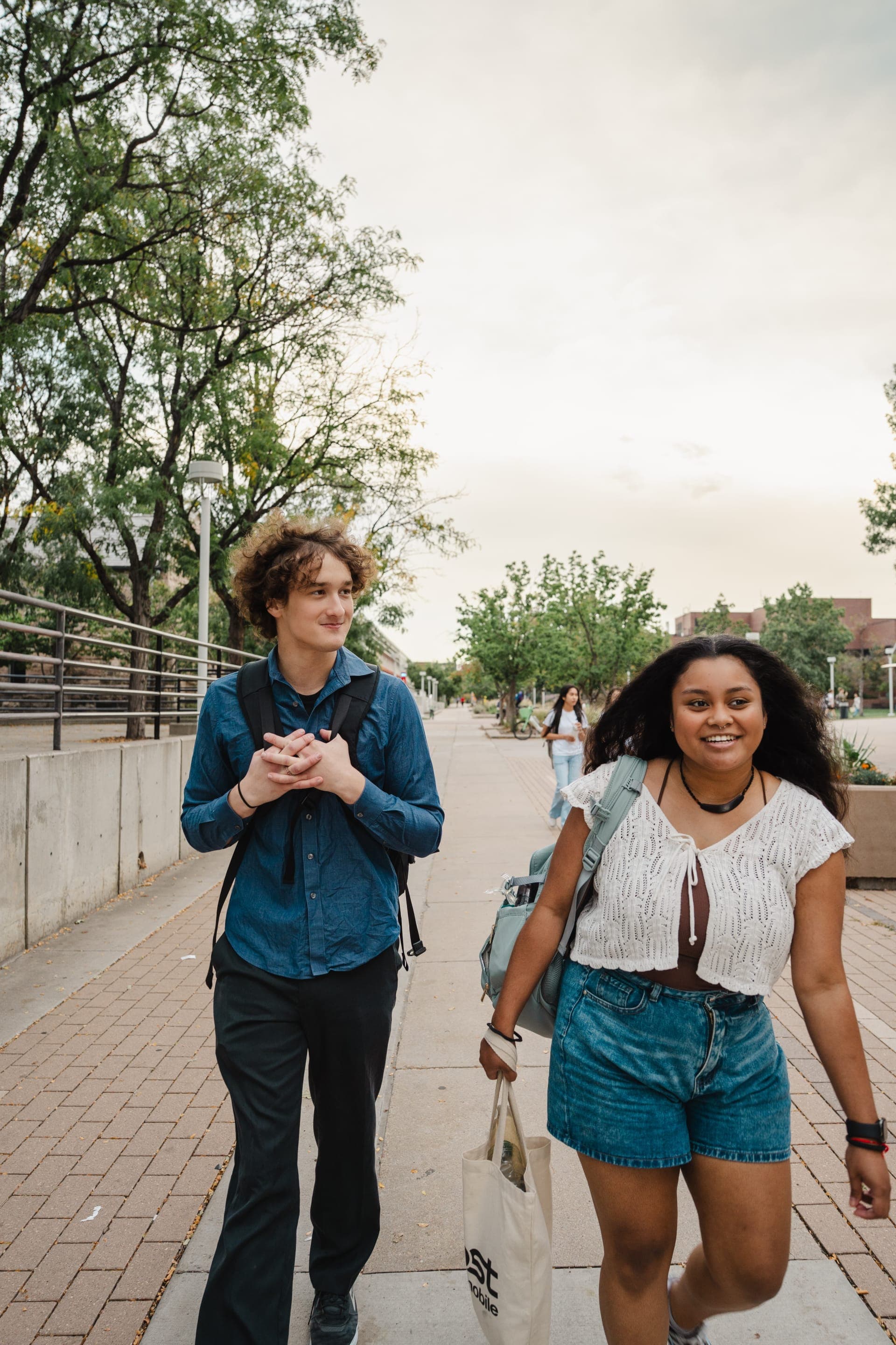 Two young people walk side by side down a tree-lined sidewalk, laughing in the evening light.