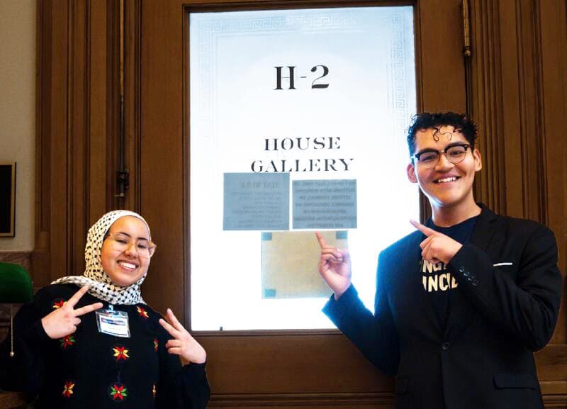 Two young advocates smile and point to the House Gallery sign inside the state capitol.